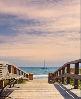 Ein Holzsteg führt an den Strand, am Horizont sieht man an Segelboot
