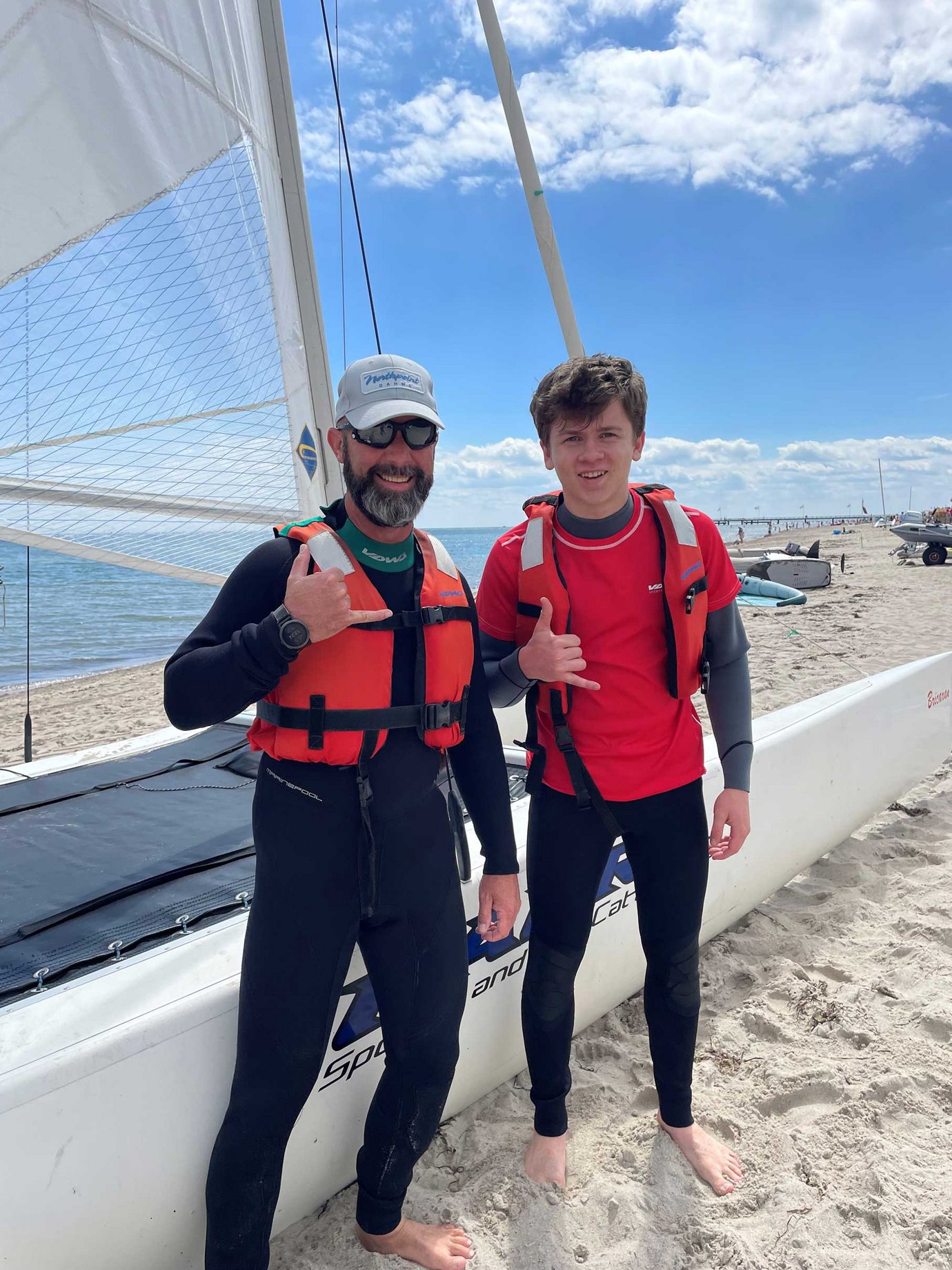 Vater und Sohn stehen bei sonnigem Wetter am Strand vor einem Katamaran und lächeln in die Kamera