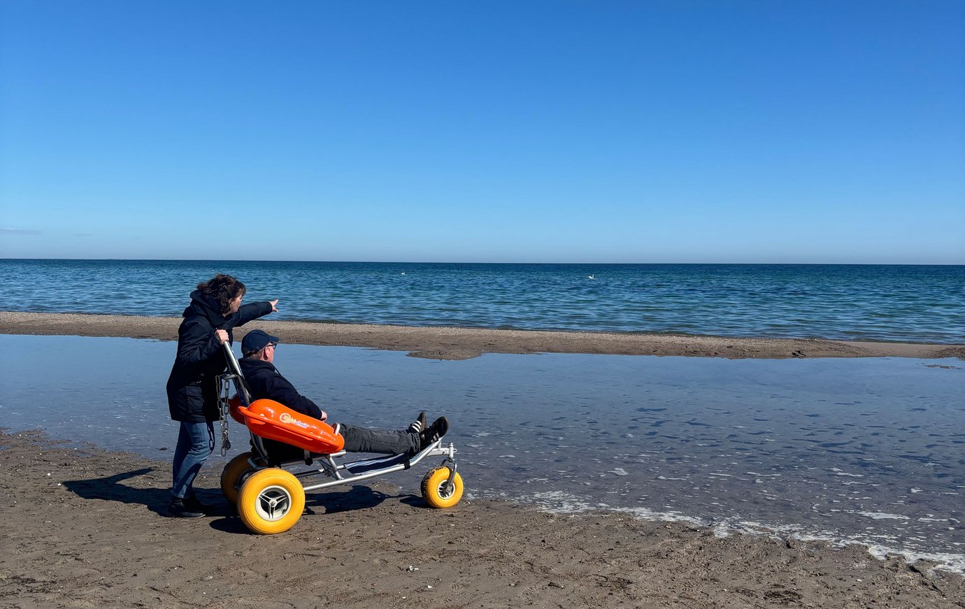 Eine Frau steht mit einem Mann im Baderollstuhl am Wasser und zeigt in die Ferne