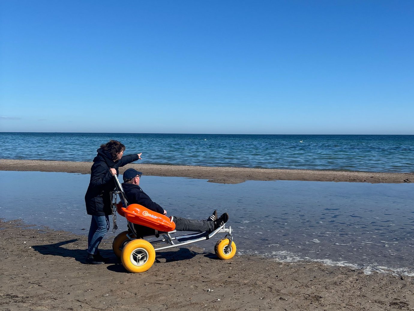 Eine Frau steht mit einem Mann im Baderollstuhl am Wasser und zeigt in die Ferne