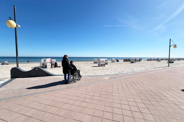 Eine Frau schiebt einen Mann im Rollstuhl bei Sonnenschein über eine Strandpromenade