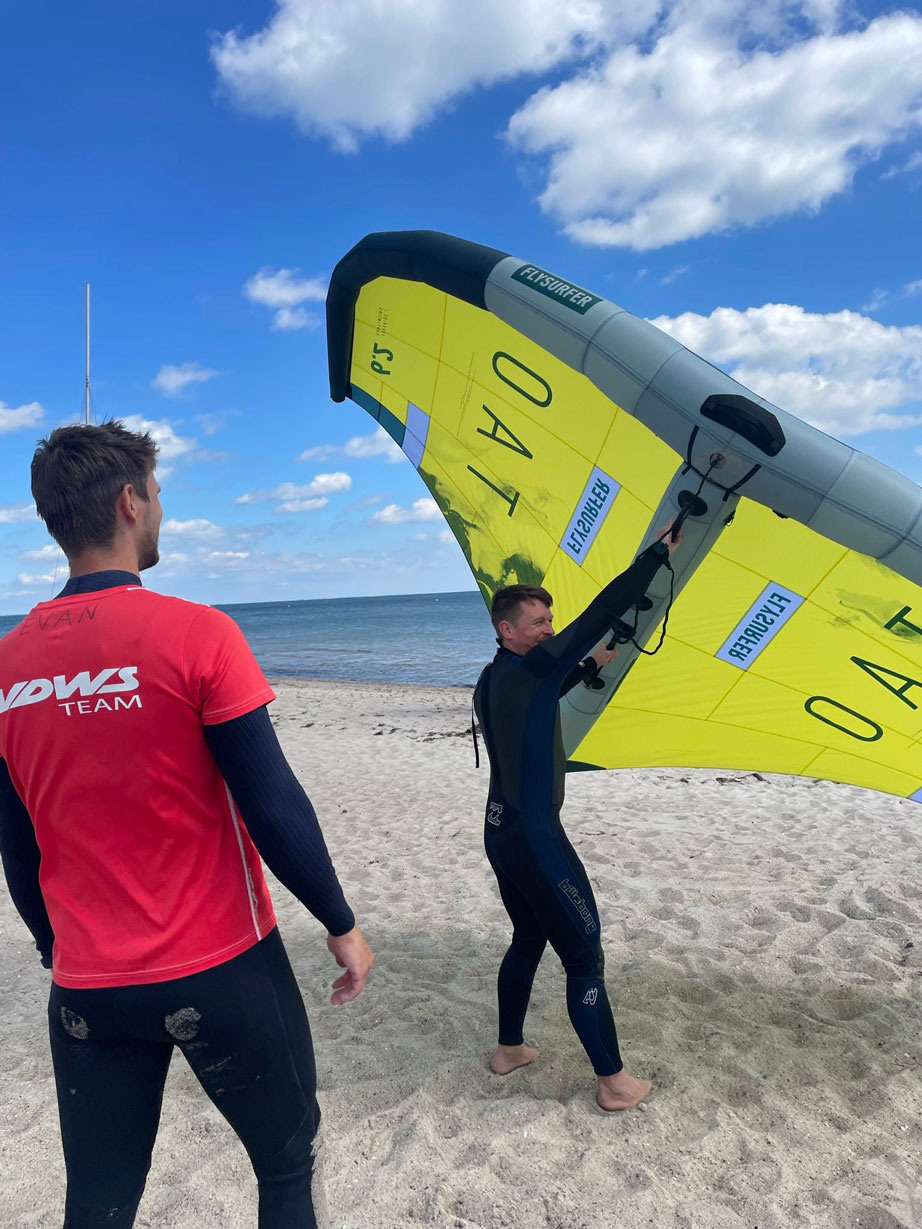 Zwei Männer stehen am Strand, einer der beiden hält das Wing-Segel